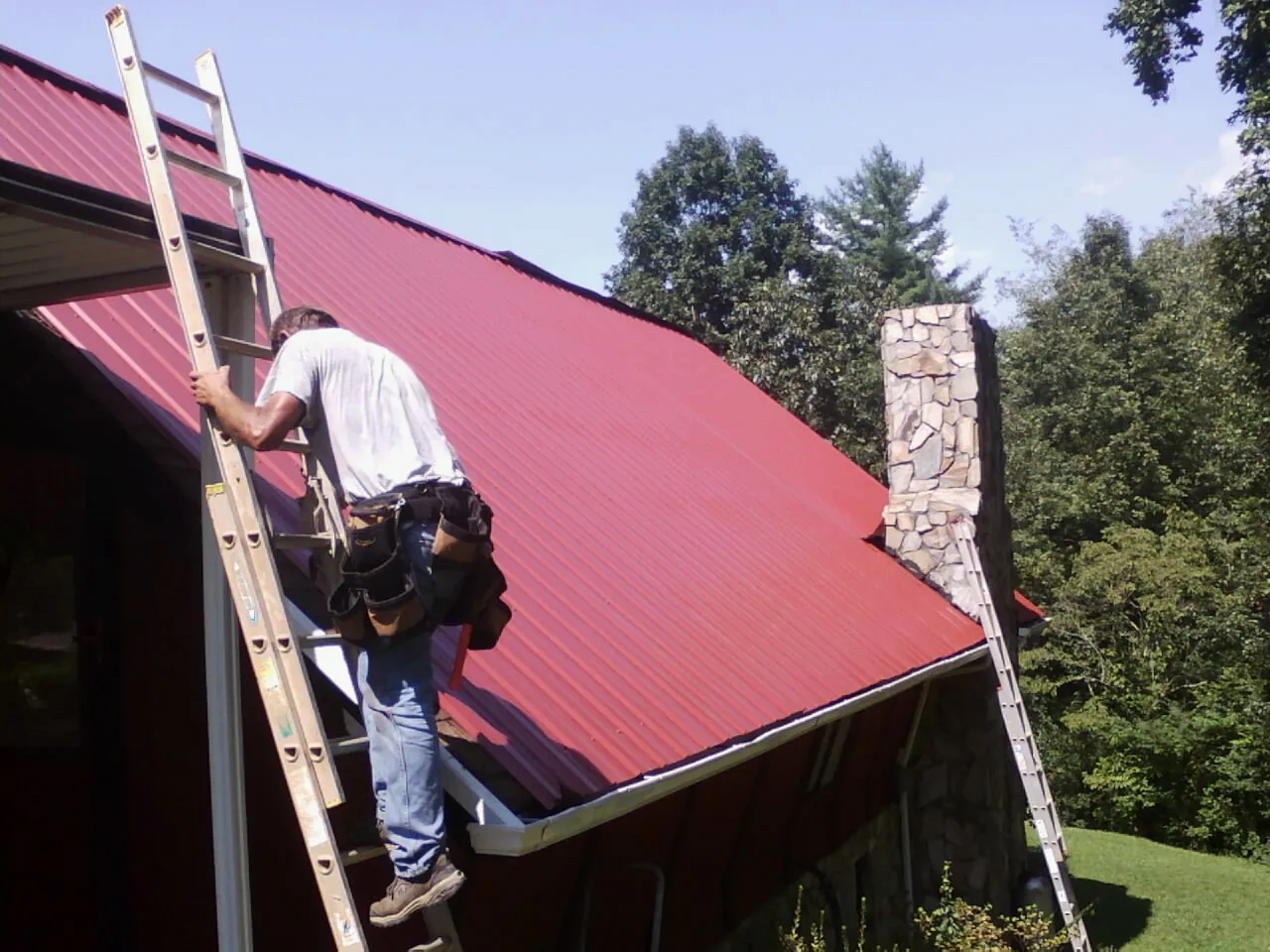 Roofer on a ladder beside a red metal roof and stone chimney — Soffit & Fascia Repair by Roofing Brothers in Wildwood